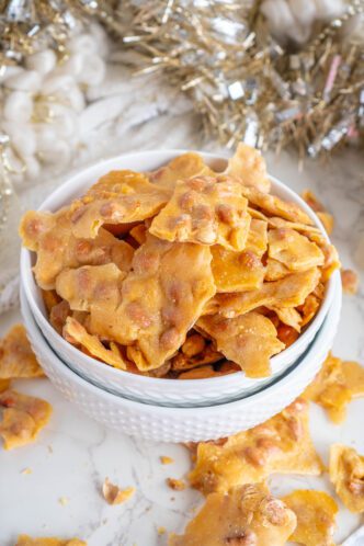 A bowl filled with homemade peanut brittle pieces, crafted from a simple microwave recipe, surrounded by festive holiday decorations.