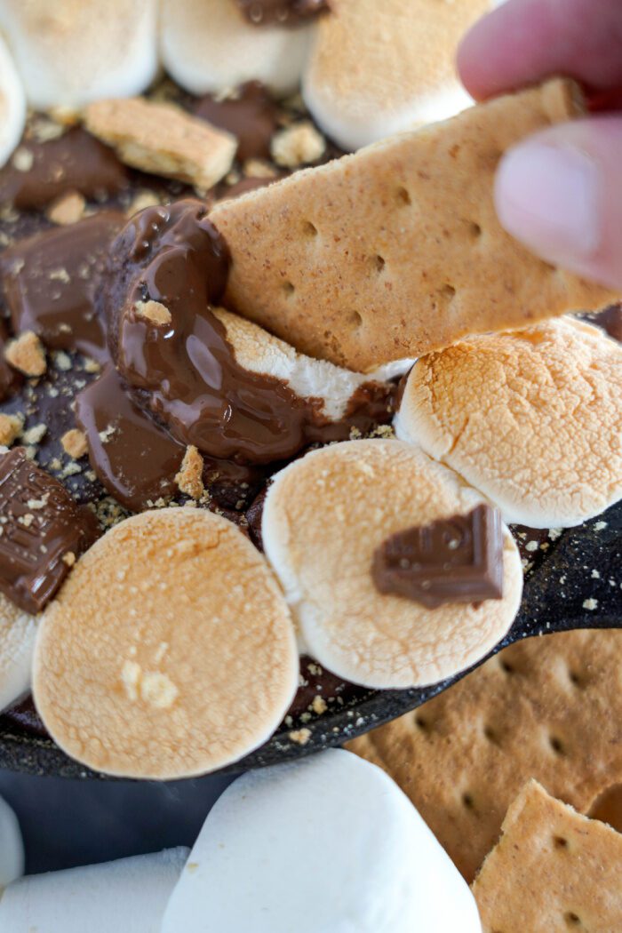 A graham cracker being dipped into a pan of melted chocolate and marshmallows.