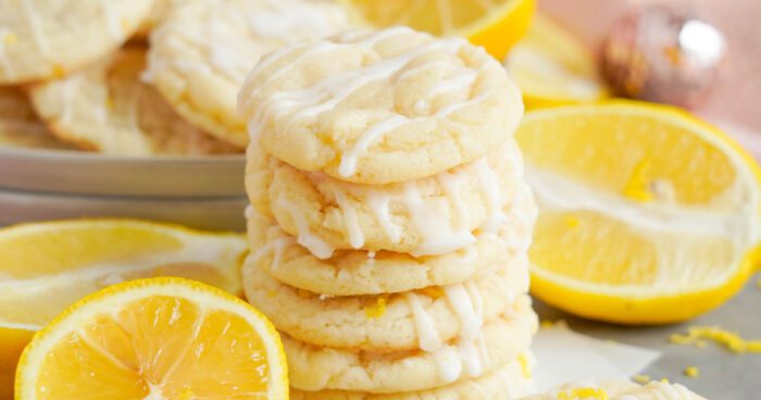 Stack of lemon sugar cookies with white glaze, surrounded by fresh lemon slices and zest on a marble countertop.