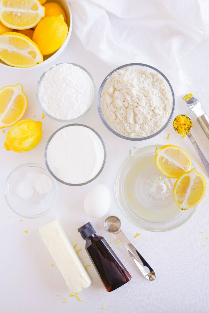 Ingredients for Lemon Sugar Cookies Recipe laid out on a white surface, including lemons, flour, sugar, butter, an egg, and vanilla extract.