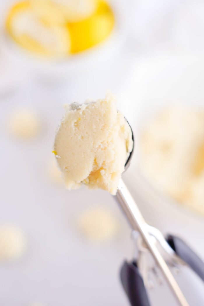 A scoop of lemon ice cream held up against a blurred background with lemon slices and a bowl of more ice cream, suggesting the flavors of a Lemon Sugar Cookies recipe.