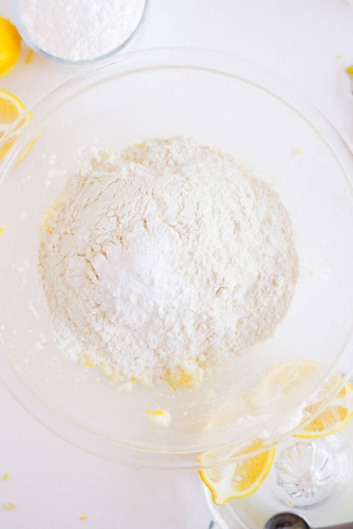 A clear glass bowl containing flour mixed with lemon zest on a white surface with lemons and baking ingredients for a Lemon Sugar Cookies recipe around it.