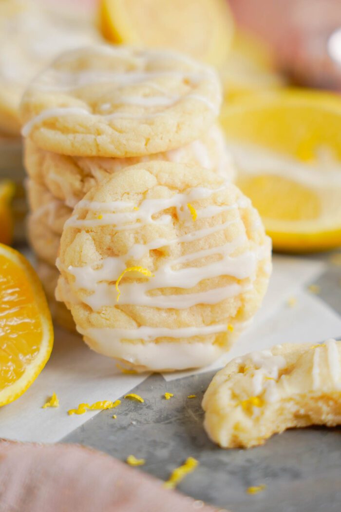 A stack of lemon sugar cookies drizzled with glaze and lemon zest on a gray surface, with fresh lemon slices beside them and a partially eaten cookie in the foreground.