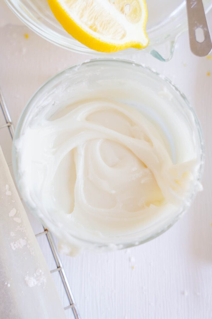 A bowl of creamy white frosting on a table beside lemon slices and a butter knife, ready for a Lemon Sugar Cookies recipe.