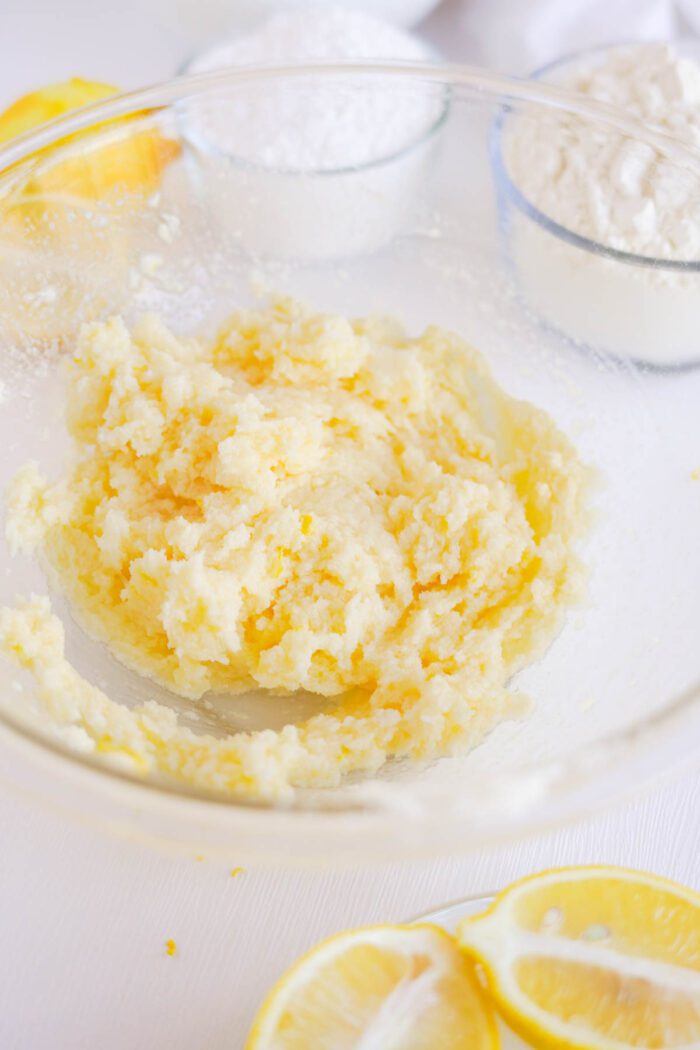 A glass bowl containing a mixture of flour and creamed butter, with sliced lemons and a sieve dusted with lemon sugar in the background.