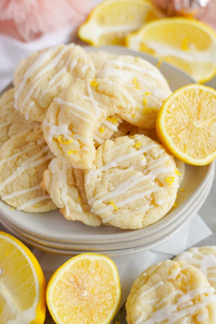 A stack of lemon sugar cookies with white glaze on a plate, surrounded by sliced lemons on a light background.