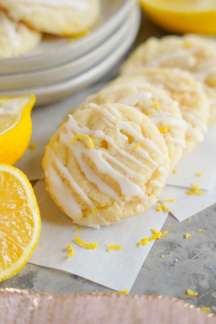 Lemon sugar cookies with white glaze on parchment paper, garnished with lemon zest, with halved lemons and a stack of cookies on a plate in the background.