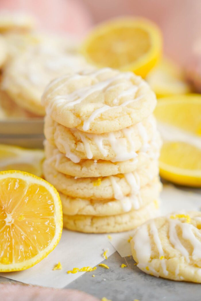 Stack of lemon sugar cookies with fresh lemon slices and zest around them on a light surface.