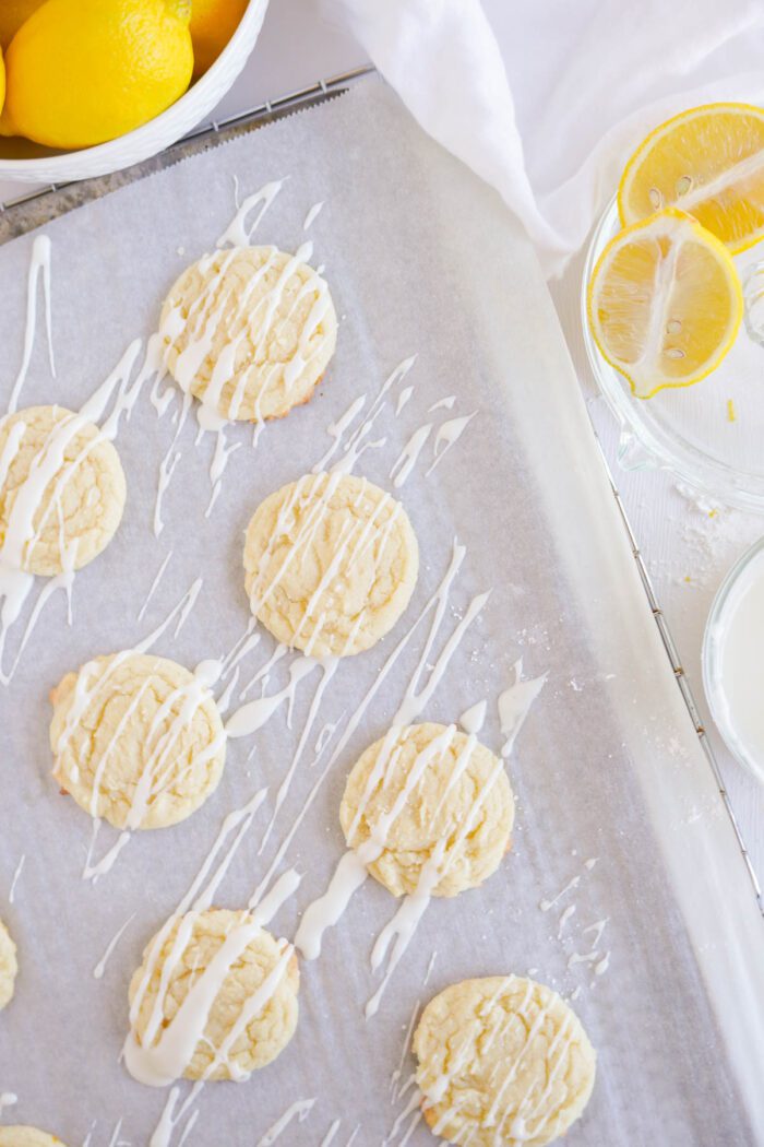 Lemon sugar cookies drizzled with white icing on parchment paper, with lemon slices and a bowl in the background.