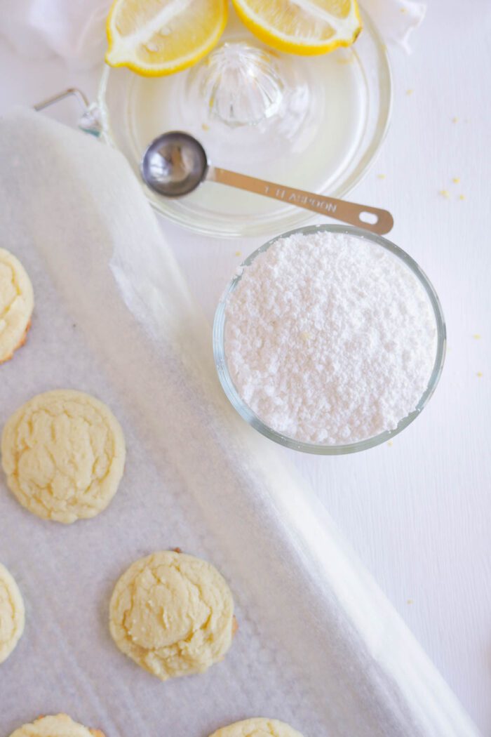 Overhead view of lemon sugar cookies on parchment paper, a bowl of powdered sugar, a plate with lemon slices, and a spoon, set on a white table.