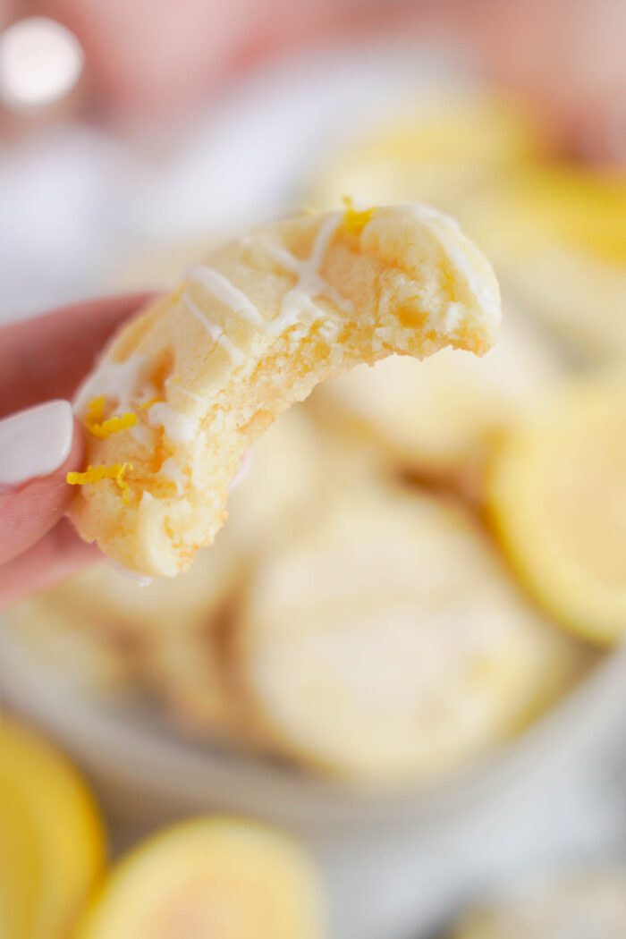 A close-up of a hand holding a bitten lemon sugar cookie with icing drizzle, with more cookies and lemon slices in the background.