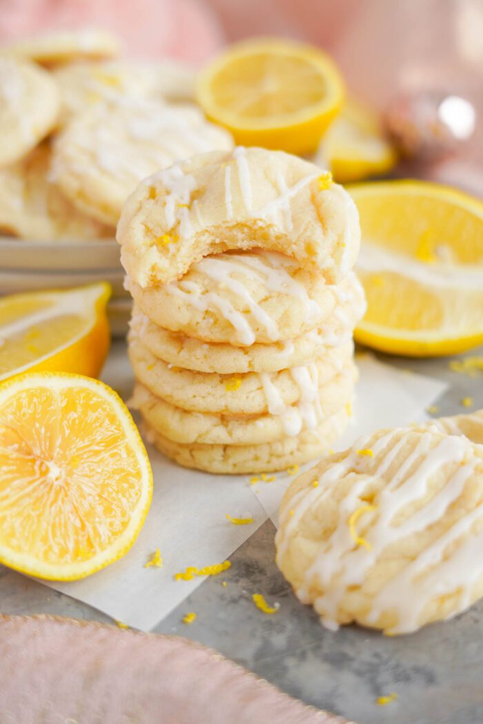 Stack of lemon sugar cookies with white icing, surrounded by fresh lemon slices on a light surface.