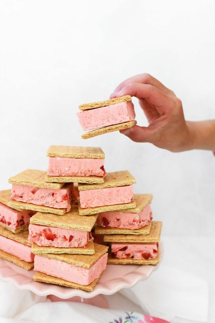 A hand holding a strawberry ice cream sandwich above a stack of ice cream sandwiches.