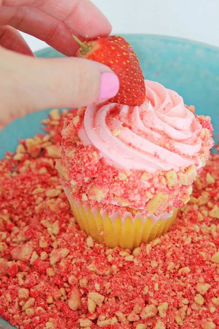 A hand adding a strawberry to a cupcake.