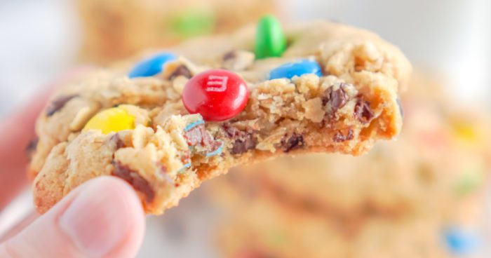 A hand holding a bitten oatmeal chocolate chip cookie with M&Ms.