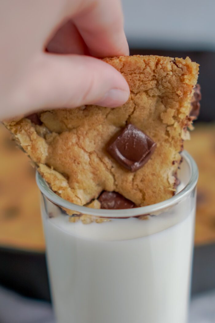 A Slice of Chocolate Chip Skillet Cookie Being Dunked Into a Glass of Milk