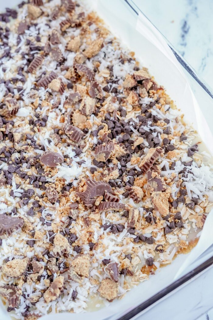 Magic bars topped with oats, chocolate chips, coconut flakes, and Reese's Cups