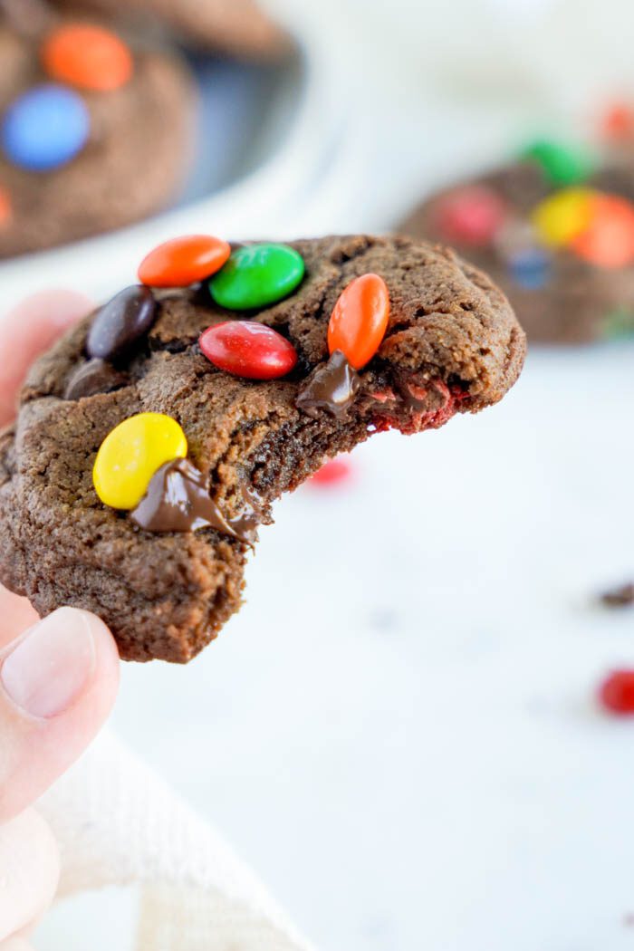 A hand holding a bitten chocolate cookie with colorful candies.