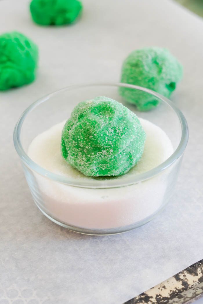 A green cookie dough ball in a glass bowl of sugar.