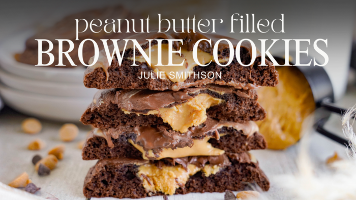 A stack of Easy Peanut Butter Brownie Cookies sits on a table, with some cookies and baking ingredients visible in the background. The text over the image reads: "Peanut Butter Filled Brownie Cookies" by Julie Smithson.