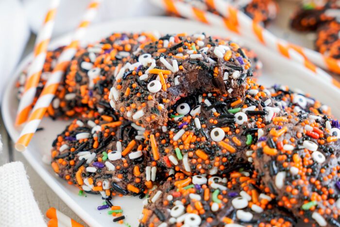 A close-up of Easy Halloween Brownie Cookies covered with orange, black, white, and purple Halloween-themed sprinkles, arranged on a white plate. One cookie has a bite taken out of it. Two striped straws are in the background.