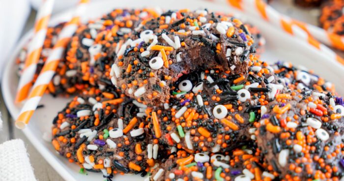 A plate of Easy Halloween Brownie Cookies decorated with colorful sprinkles, including white candy eyes. One cookie has a bite taken out of it. Straw-striped orange and white paper straws are in the background.