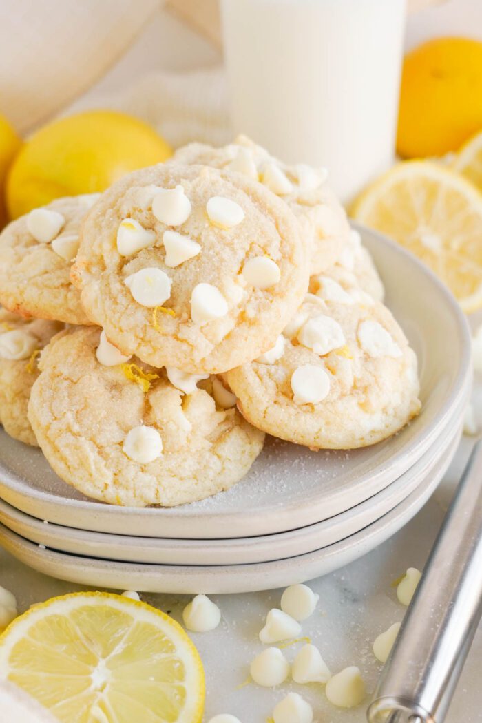 A stack of Lemon White Chocolate Chip Cookies on a plate, accompanied by fresh lemons and a glass of milk.