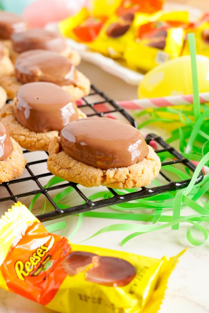 Peanut butter cookies topped with Reese's chocolate candy on a cooling rack, with candy wrappers in the foreground.