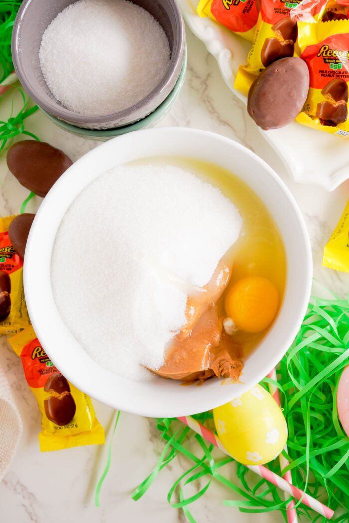 Ingredients for baking Reese's Egg Peanut Butter Cookies for Easter laid out on a counter, including eggs, sugar, peanut butter cups, and decorative grass.