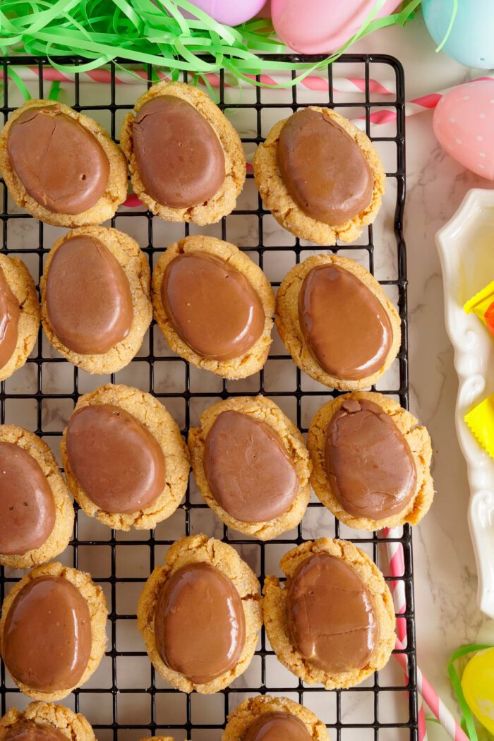 Freshly baked Reese's Egg Peanut Butter cookies with a chocolate topping cooling on a wire rack, surrounded by Easter decorations.