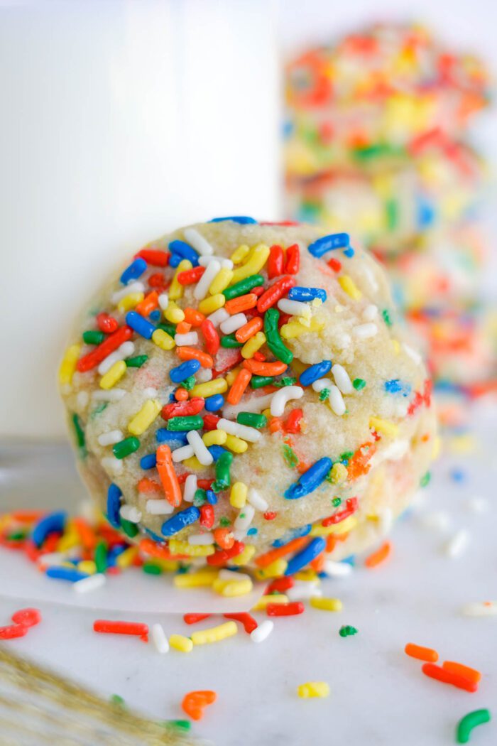 Sugar cookie with rainbow sprinkles in focus, with a glass of milk and additional sprinkles in the background.