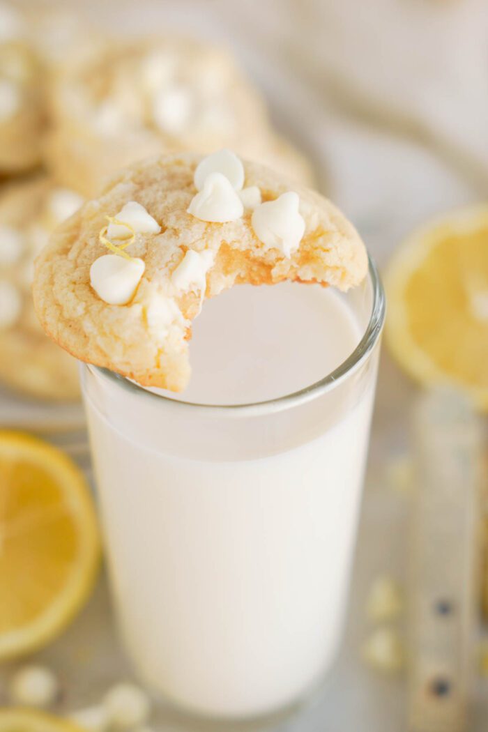 A lemon white chocolate chip cookie with a bite taken out of it balanced on a glass of milk, with lemons and cookies in the background.