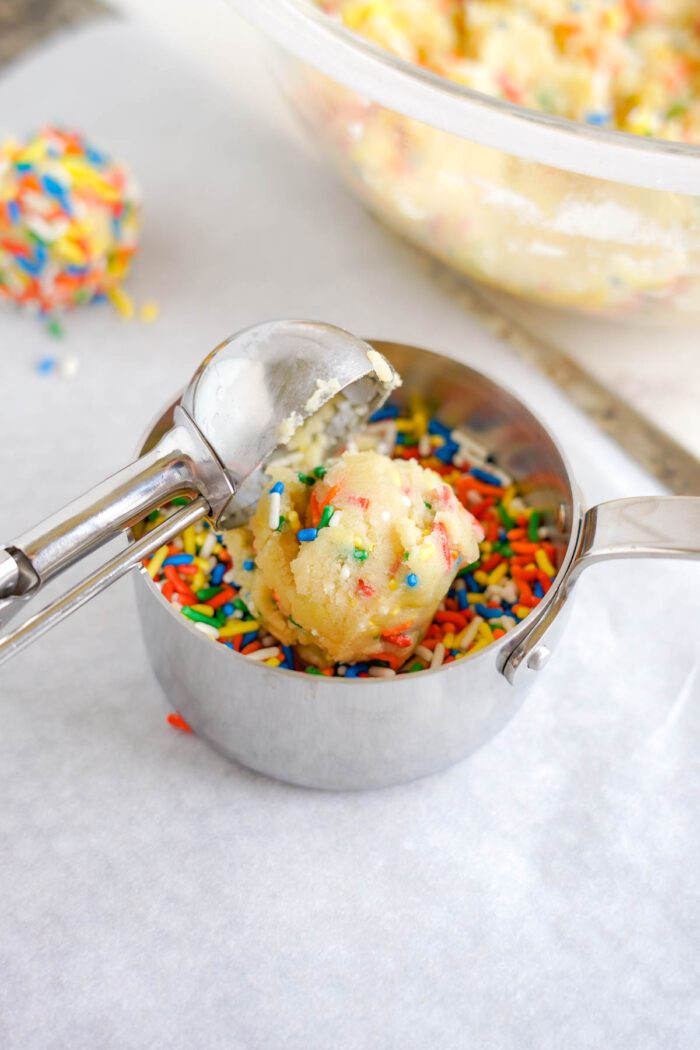 A scoop of cookie dough with sprinkles rests on a scooper above a bowl and surface scattered with colorful sprinkles.