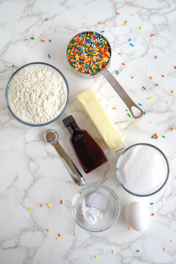 Ingredients for baking laid out on a marble countertop, including flour, sprinkles, butter, vanilla extract, sugar, baking powder, and an egg.