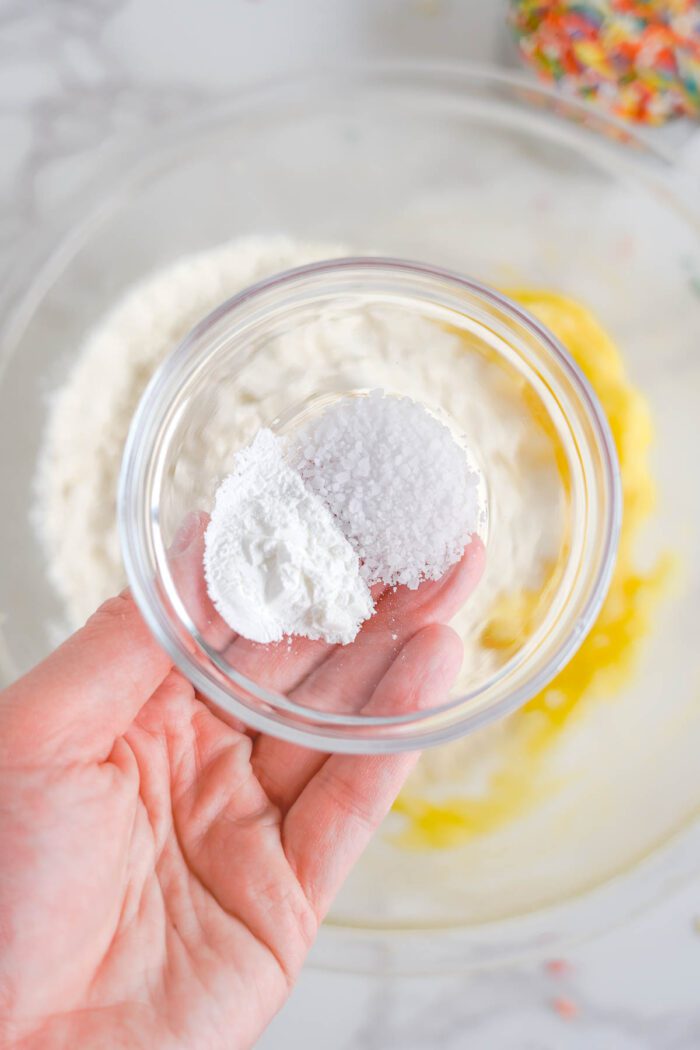 A person holding a small glass bowl containing baking powder over a larger bowl with a mix of eggs and flour.