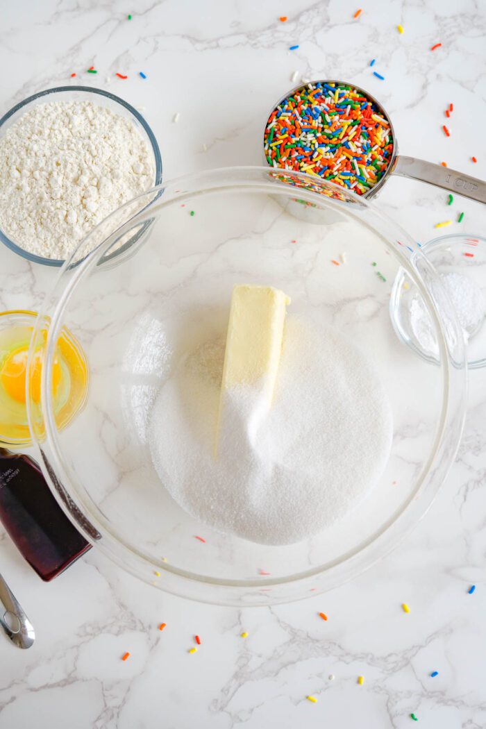 Ingredients for baking arranged on a countertop, including flour, sugar, butter, eggs, vanilla extract, and sprinkles.