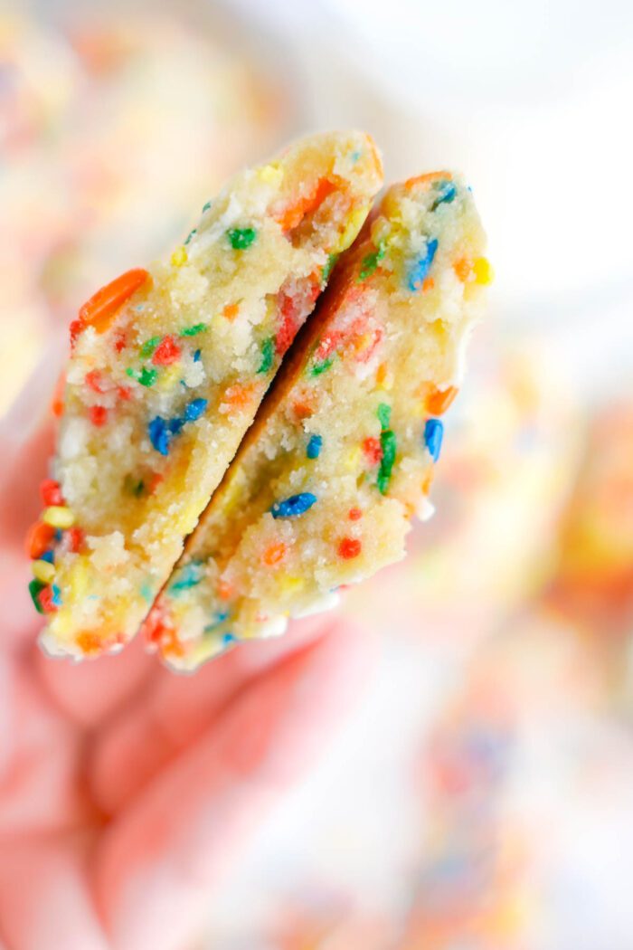 A hand holding a split funfetti cookie showing colorful sprinkles inside.