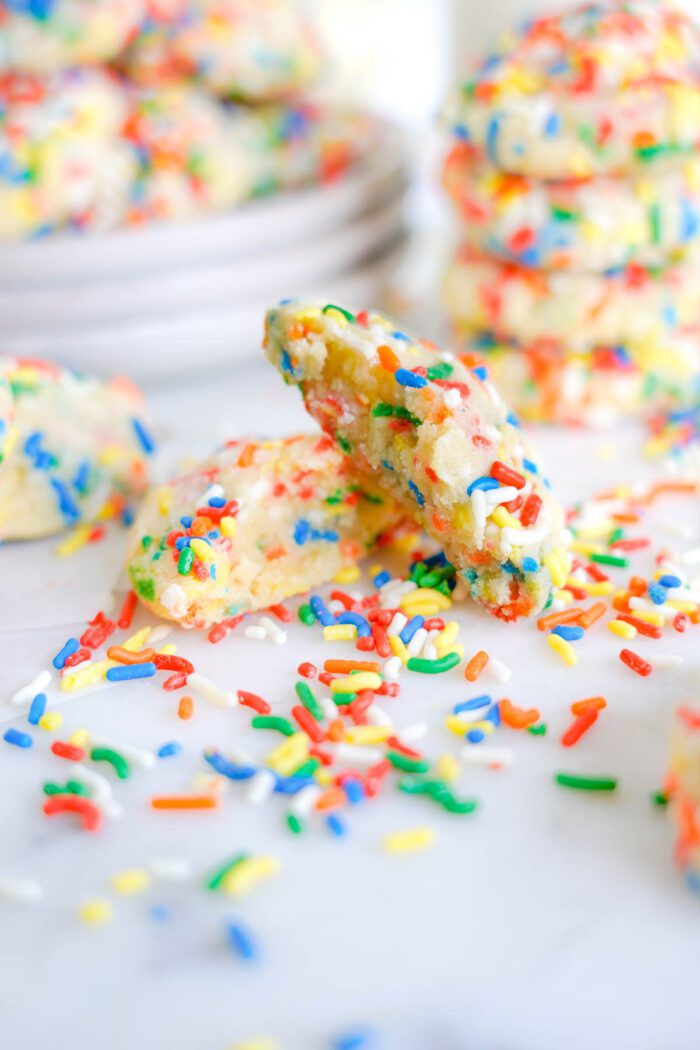 Stack of freshly baked sprinkle cookies with one cookie broken in half in front, showcasing the soft interior.