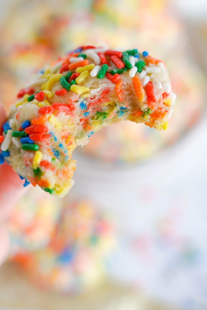 A close-up of a hand holding a bitten funfetti sugar cookie with colorful sprinkles.