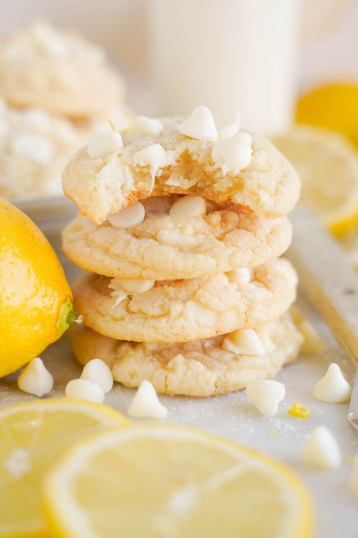 A stack of Lemon White Chocolate Chip Cookies garnished with fresh lemon slices and a glass of milk in the background.