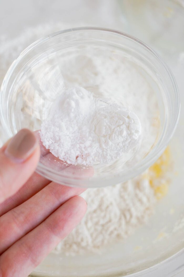 A person holding a small clear bowl containing a scoop of lemon white chocolate chip cookie powder above a larger bowl with other ingredients.