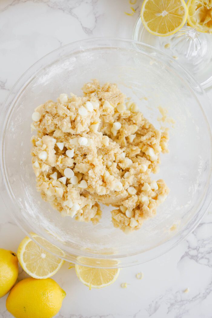 A bowl of crumbly Lemon White Chocolate Chip Cookie dough, next to fresh lemons on a marble surface.