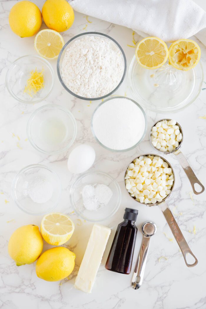 Ingredients for baking Lemon White Chocolate Chip Cookies arranged neatly on a marble countertop, including lemons, flour, sugar, eggs, and butter.
