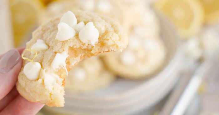 A close-up view of a hand holding a lemon white chocolate chip cookie with a bite taken out, with more cookies in the background.