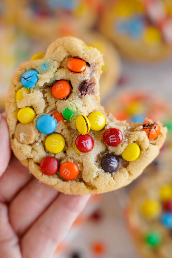 A hand holding a colorful peanut butter M&M cookie with a bite taken out of it.