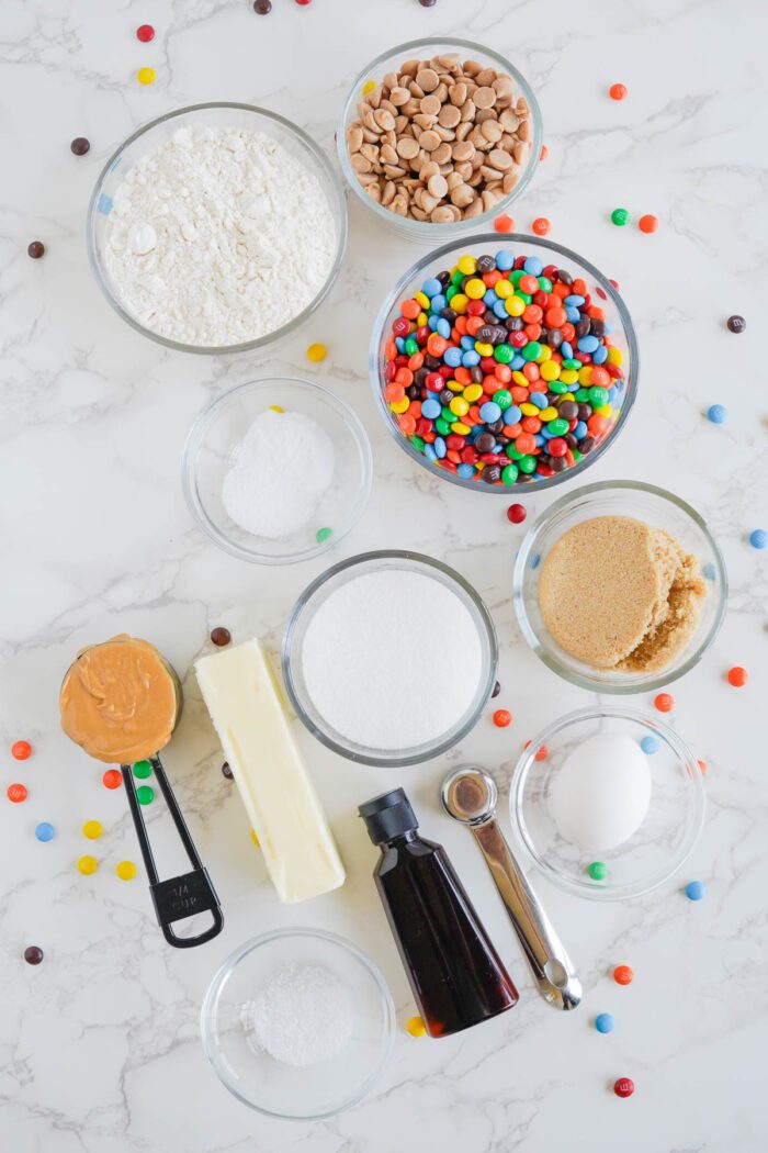 Ingredients for the Best Peanut Butter M&M Cookies Recipe laid out on a marble countertop, including flour, chocolate chips, M&M candies, peanut butter, butter, sugars, an egg, baking powder