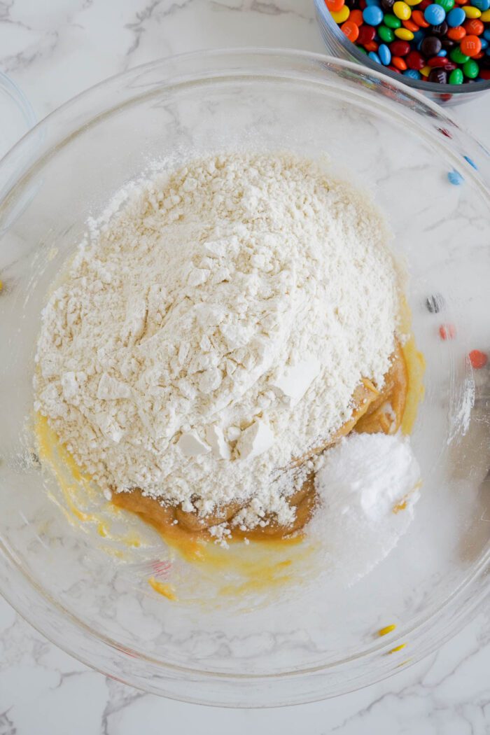 A glass bowl containing flour and a liquid mixture on a kitchen counter with colorful Peanut Butter M&M candies scattered around.