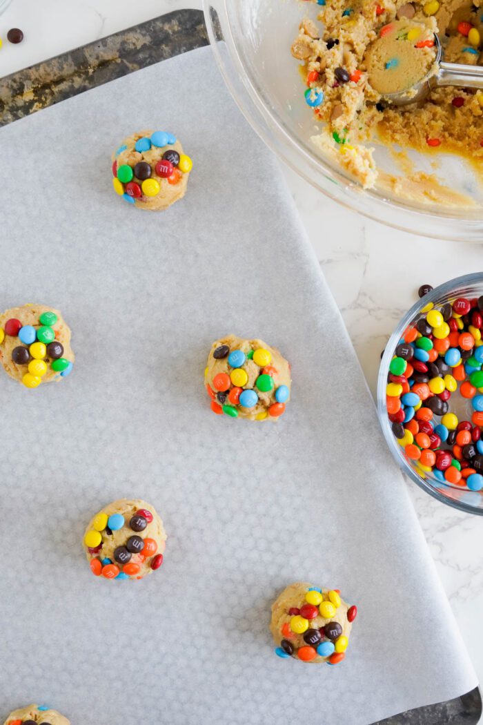 Best Peanut Butter M&M Cookies dough balls with colorful candy pieces are placed on parchment paper, ready for baking, with additional candy and dough in bowls beside them.