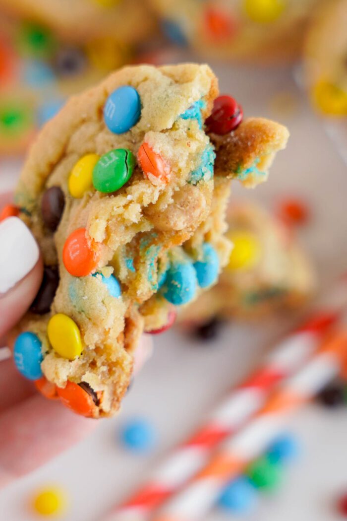 A close-up of a hand holding a colorful, best peanut butter M&M candy-studded cookie broken in half, revealing a soft and chewy texture inside.