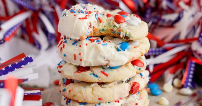 A stack of four Easy 4th of July Cake Mix Cookies with white frosting, red and blue sprinkles, and red and blue candies, surrounded by festive red, white, and blue party decorations. The top cookie has a bite taken out.